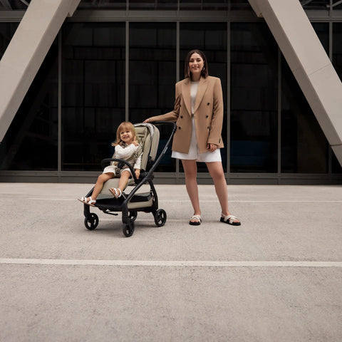A woman in a beige blazer and white skirt stands next to a taupe-colored Redsbaby Aeron stroller with a black frame. The stroller is holding a smiling child dressed in white, sitting comfortably with their feet resting on the footrest. The background features a modern building with large windows and geometric architecture.