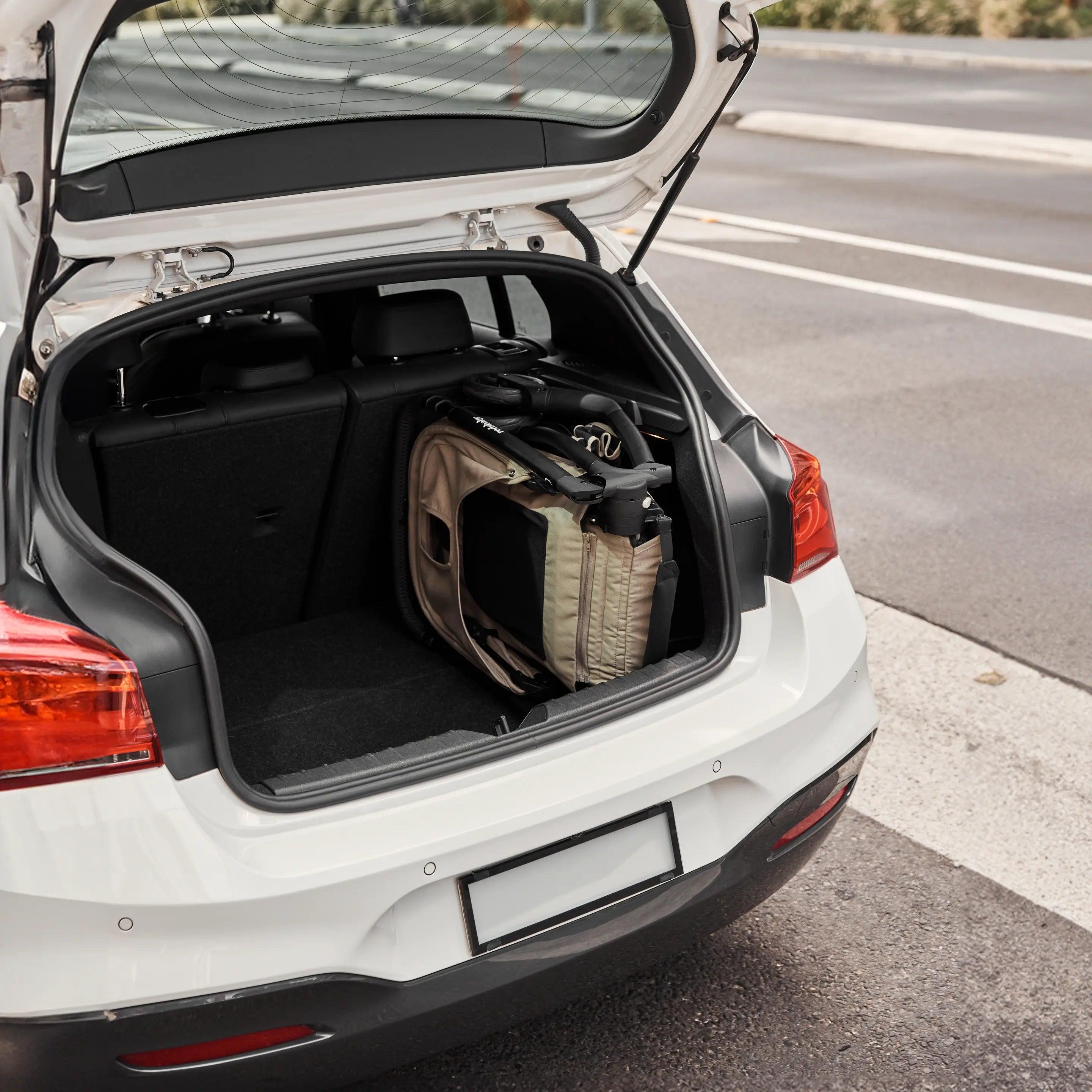 A folded Redsbaby Aeron stroller stored neatly in the trunk of a white car. The stroller is compactly folded, showcasing its portability and convenience for travel. The car is parked on a road with a blurred background of greenery.