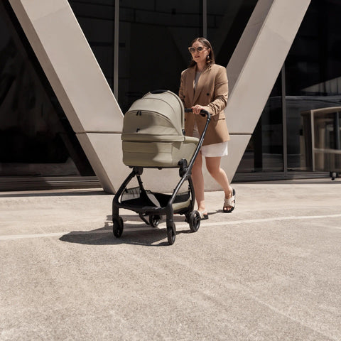 A woman in a beige blazer and white skirt walks outside, pushing a taupe-colored Redsbaby Aeron stroller with a black frame. The stroller features a canopy and spacious storage basket underneath. The background includes a modern building with geometric architecture and large windows.