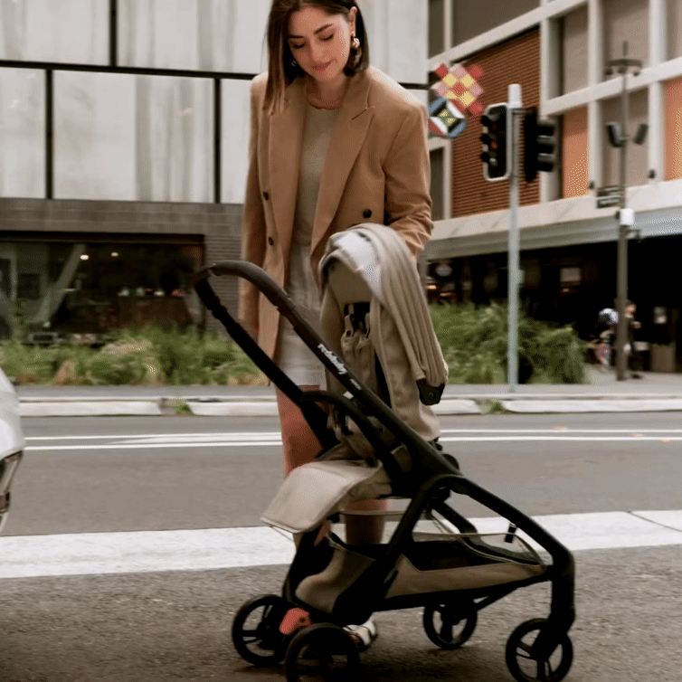 A woman in a beige blazer and white skirt is folding a taupe-colored Redsbaby Aeron stroller with a black frame on a city street. The background features urban buildings, traffic lights, and greenery, showcasing the stroller's ease of use and portability in an urban environment.