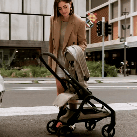 A woman in a beige blazer and white skirt is folding a taupe-colored Redsbaby Aeron stroller with a black frame on a city street. The background features urban buildings, traffic lights, and greenery, showcasing the stroller's ease of use and portability in an urban environment.